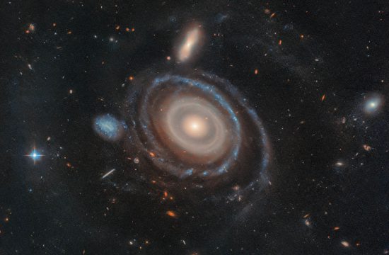 A large galaxy is at center, and a significantly smaller galaxy is to its left. The large galaxy, nicknamed the Bullseye, is mostly face-on, but the top appears slightly tilted away. It has several rings. Its circular core is bright white at the very center, but light yellow overall. Going outward, there are gaps between the rings. The core is surrounded by two slightly lighter yellow rings, which also appear to be overlapping. The next ring is slightly more transparent and yellow. The two or three rings that are farther out are bluer, sometimes with blue clumps. The widest ring is also blue, but also the most transparent. At 9 o’clock is a small dwarf galaxy. It is about the same size as the yellow core of the Bullseye. The dwarf galaxy is blue, with many dots. It looks like the edge of the Bullseye might touch the dwarf galaxy. Both galaxies are set on the black background of space, which is dotted with a range of galaxies in different shapes, colors, and sizes, along with one foreground star at left.